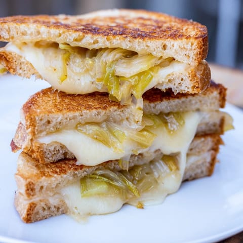 The Havarti & Caramelized Leek Grilled Cheese is sliced in half on a rustic wooden board, revealing its gooey, savory filling.