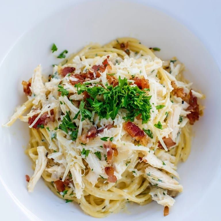 Close-up of Chicken Carbonara, highlighting glossy sauce-coated noodles, crispy bacon bits, and a generous dusting of grated Parmesan cheese.
